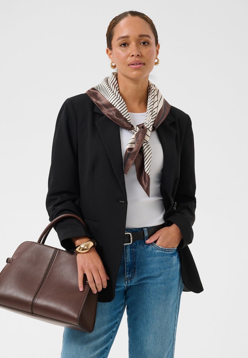 Femme portant un blazer noir, un haut blanc, une écharpe en soie rayée, un jean bleu, tenant un sac à main en cuir marron et portant des boucles d’oreilles créoles en or et un bracelet.