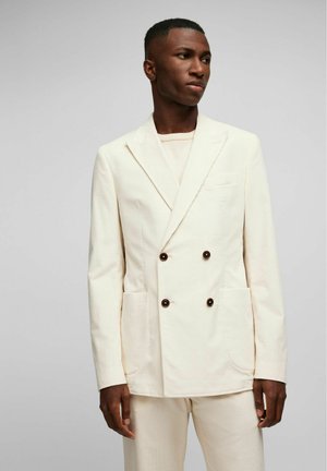 Crème kleurige dubbelzijdige blazer met gestructureerde stof, bruine knopen en opgestikte zakken, gecombineerd met een bijpassend top van een glad materiaal.