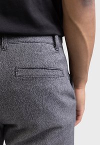 Pantalon gris texturé avec une poche arrière bordée de coutures et une ceinture plate. Porté avec une chemise noire.