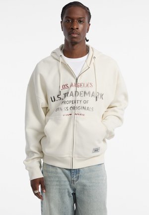 Crème rits hoodie van een getextureerde stof, met vooraan branding in rood en grijs, en een klein label aan de onderkant. Draag ik over een wit shirt.