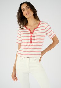 Top de manga corta a rayas en color crema con líneas horizontales rojas, con escote redondo y botonera. Combinado con jeans blancos.