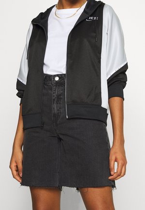 Personne portant un sweat à capuche zippé noir et blanc, un t-shirt blanc, une chaîne en or et une jupe en jean noire à ourlet brut.