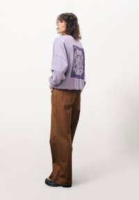 Helllila Sweatshirt mit einem dunkel-lila Aufdruck auf der Rückseite, kombiniert mit braunen Weitbein-Hosen und schwarzen Plateausandalen.