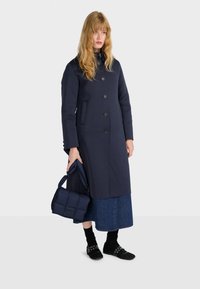 Cappotto lungo in misto lana blu navy con chiusura a bottoni, tasche laterali, una forma aderente, abbinato a una borsa trapuntata blu navy e calzature nere.