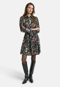 Blumenhemdkleid in Dunkelblau, mit Kragen, langen Ärmeln und einem gebundenen Taillengürtel. Getragen mit kniehohen schwarzen Stiefeln.