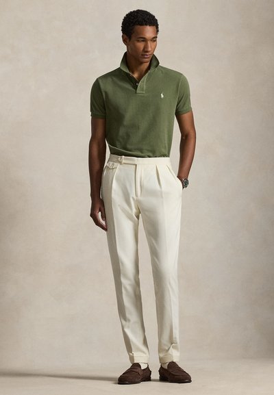 Vīrietis valkā olīvu zaļu polo krekliņu ar paceltu apkakli, krēmīgas krāsas krokotās bikses, brūnas mokasīnus un pulksteni, stāvot ar vienu roku kabatā.