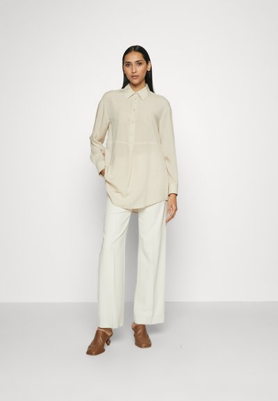 Camicia beige a maniche lunghe con bottoni e vestibilità comoda, abbinata a pantaloni bianchi a gamba larga e mule marroni. Texture liscia, design minimale.