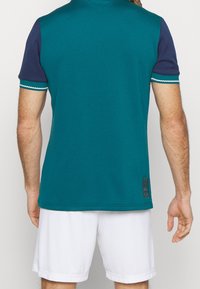 Polo bleu sarcelle avec manches bleu marine, présentant des accents en zigzag blancs aux poignets, conçu avec une texture lisse et un écusson logo à l'ourlet.