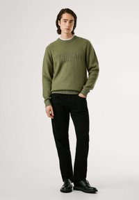 Žalias raukšlėtas megztinis su įspaustu logotipu, derinamas su juodomis tiesiomis kelnėmis ir juodais bateliais su raišteliais ant paprasto fono.