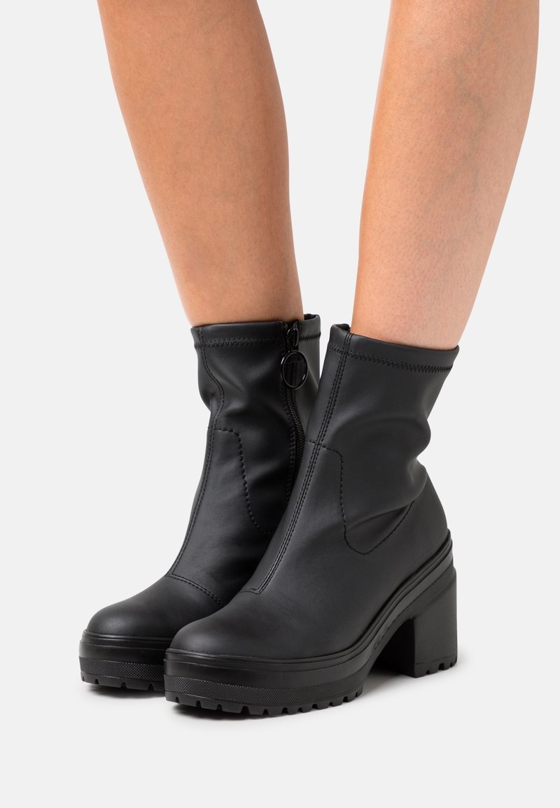 Bottes mi-mollet en cuir noir avec fermetures éclair latérales, talons épais et semelles en caoutchouc épaisses, portées sur jambes nues sur un fond clair.