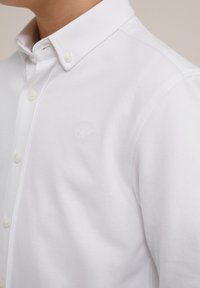 Man klädd i en vit skjorta med knapp och med knäppt krage samt ett diskret broderat logo på bröstet.