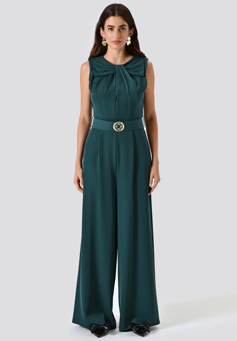 Teal Jumpsuit mit einem gefältelten Oberteil, einem taillierten Gürtel mit einer dekorativen Schnalle und weit fließenden Beinen. Tragend mit schwarzen Schuhen mit Schleifenakzenten.