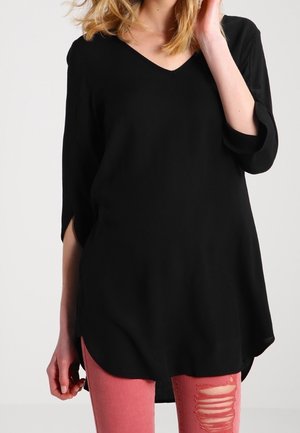 Kvinde iført en løs sort tunika med V-udskæring og 3/4 ærmer samt rosa slidte skinny jeans, stående foran en ensfarvet baggrund.