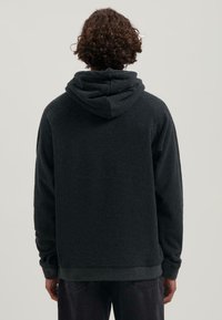 Persona con capelli ricci che indossa una felpa con cappuccio grigio scuro e pantaloni neri, in piedi di spalle contro uno sfondo chiaro e uniforme.