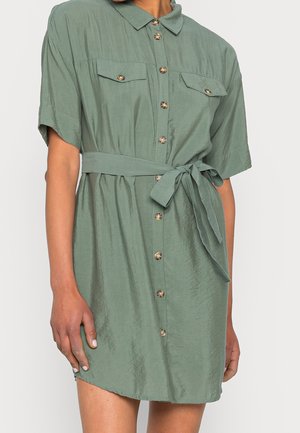 Vihreä, lyhythihainen paitamekko, jossa on etunapit, rintataskut ja solmittava vyötärövyö, pukeutuneena tummaihoiselle henkilölle.