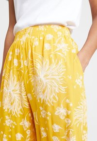 Gula blommiga vida byxor med elastiskt midjeband, med vita botaniska mönster och sidofickor. Mjuk, lätt tyg.