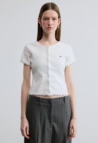 Λευκό ριπέν κοντό τοπ με κυματιστά άκρα, πέντε κουμπιά μπροστά και μικρό λογότυπο. Συνδυασμένο με γκρι παντελόνι με λεπτές ρίγες.