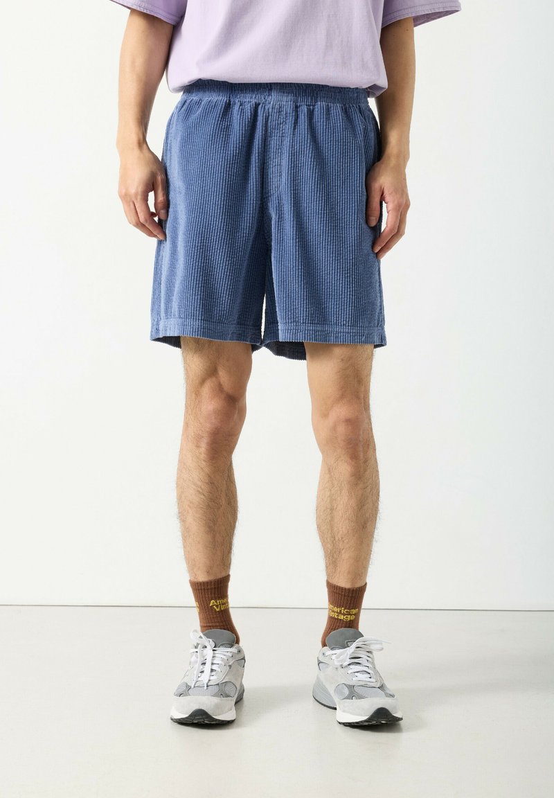 Mann trägt blaue, strukturierte Shorts, braune Socken mit gelbem Text, weiße und graue Turnschuhe sowie ein lavendelfarbenes Hemd und steht auf einem weißen Boden.