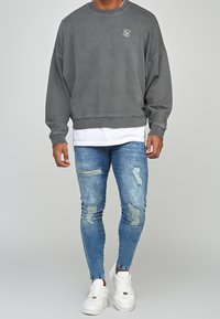 Sudadera gris de gran tamaño, camisa blanca en capas por debajo, jeans ajustados azules desgastados y zapatillas blancas con un destacado detalle de costura.