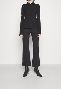 Schwarzes Langarmhemd mit Kragen und Knöpfen, kombiniert mit schwarzen Schlaghosen. Beide Kleidungsstücke verfügen über eine glatte Textur.