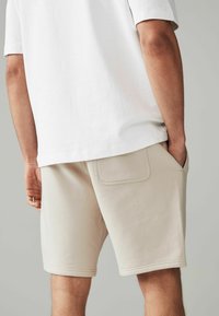 Shorts en coton beige avec une poche arrière, ornés de détails de couture et d'une coupe décontractée, associés à un top blanc à manches courtes.