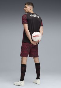 Equipación de fútbol del RB Leipzig en negro y burdeos con pantalones cortos estampados, botas blancas y calcetas negras, sosteniendo un balón de fútbol blanco con acentos rojos.