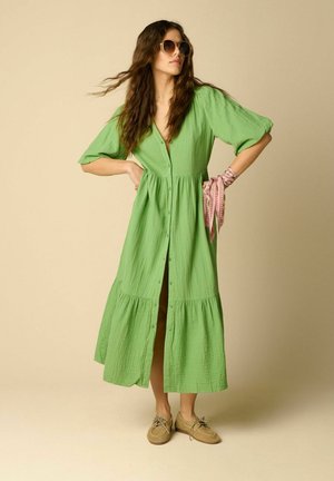 Femme portant une longue robe verte boutonnée, un foulard rose à motifs au poignet, des chaussures beiges et des lunettes de soleil rondes, debout les mains sur les hanches.