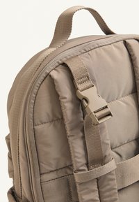 Bežový vystužený batoh s textúrovaným povrchom, nastaviteľnými popruhmi a plastovou sponou. Zipsový vrch s vystuženými švami.