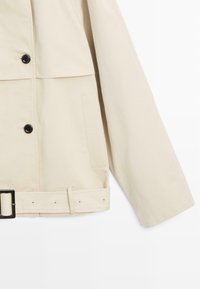 Giacca beige con bottoni neri, una tasca rettangolare sul lato e una cintura coordinata con una fibbia rettangolare nera.