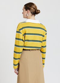Gelbes Langarmoberteil mit vertikalen grünen und weißen Streifen sowie einem weißen Kragen. Dazu wird ein beigefarbener Rock getragen; glatte Textur.