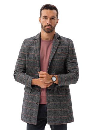 Blazer grigio a scacchi con accenti rossi, vestibilità su misura, rever a punta e due bottoni. Indossato sopra un pullover rosa chiaro, abbinato a un orologio.