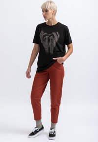Zwarte katoenen t-shirt met een grafische print van handen; gecombineerd met roestkleurige broeken, weergegeven op een model dat zwarte sneakers draagt.