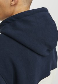 Sudadera azul marino con tejido texturizado, que presenta una gran capucha y una banda blanca en contraste en la parte inferior. Diseño simple sin acentos adicionales.