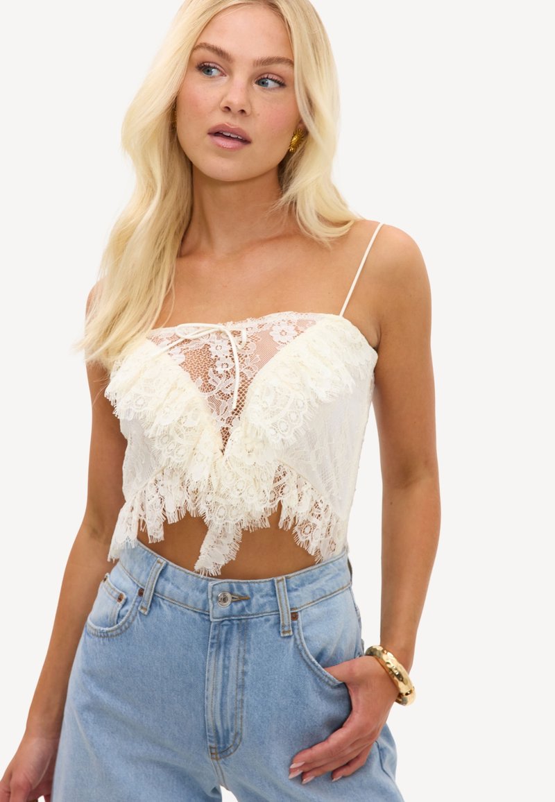 Femme blonde portant un caraco court en dentelle blanche à fines bretelles et un jean taille haute bleu clair, posant avec une main dans la poche.