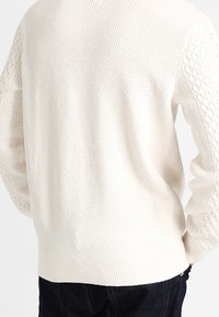 Pull en maille texturée blanche avec motifs torsadés sur les manches, porté avec un pantalon sombre, vu de dos.
