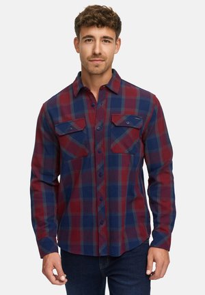 Mann in einem langärmligen rot-blauen karierten Hemd mit Knopfverschluss und Fronttaschen, steht vor einem weißen Hintergrund.
