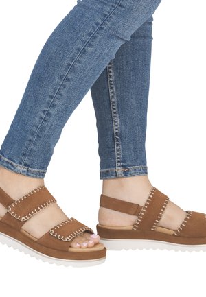 Pieds portant des sandales en daim marron avec des coutures blanches et des semelles blanches épaisses, assorties à un jean skinny bleu.