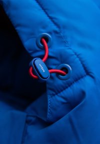 Close-up van een blauw jack met een rood trekkoord door blauwe oogjes heen en vastgezet met een blauwe plastic stopper.