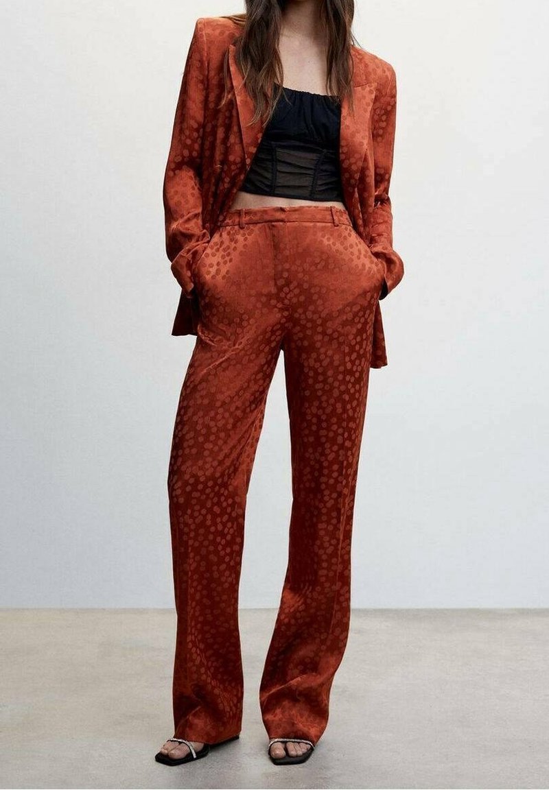 Femme portant un costume à motifs orange brûlé avec un pantalon ample et un blazer par-dessus un top court noir et des sandales noires. Mains dans les poches.