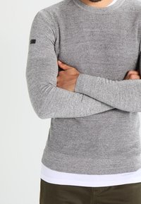 Maglione grigio a maniche lunghe in maglia con una finitura testurizzata, collo rotondo e una vita liscia, dotato di un piccolo etichetta nera sul braccio sinistro.