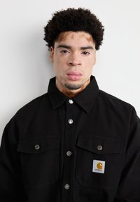 Sort lærredjakke med sølvfarvede trykknapper, to brystlommer og et Carhartt-logo patch i gul og hvid.