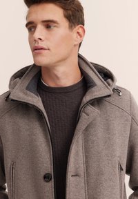 Grau melierter Woll-Mix-Jacke mit einem Reißverschluss und Knopfverschluss, mit Kapuze, seitlichen Taschen und glatter Textur. Über einem strukturierten braunen Pullover getragen.
