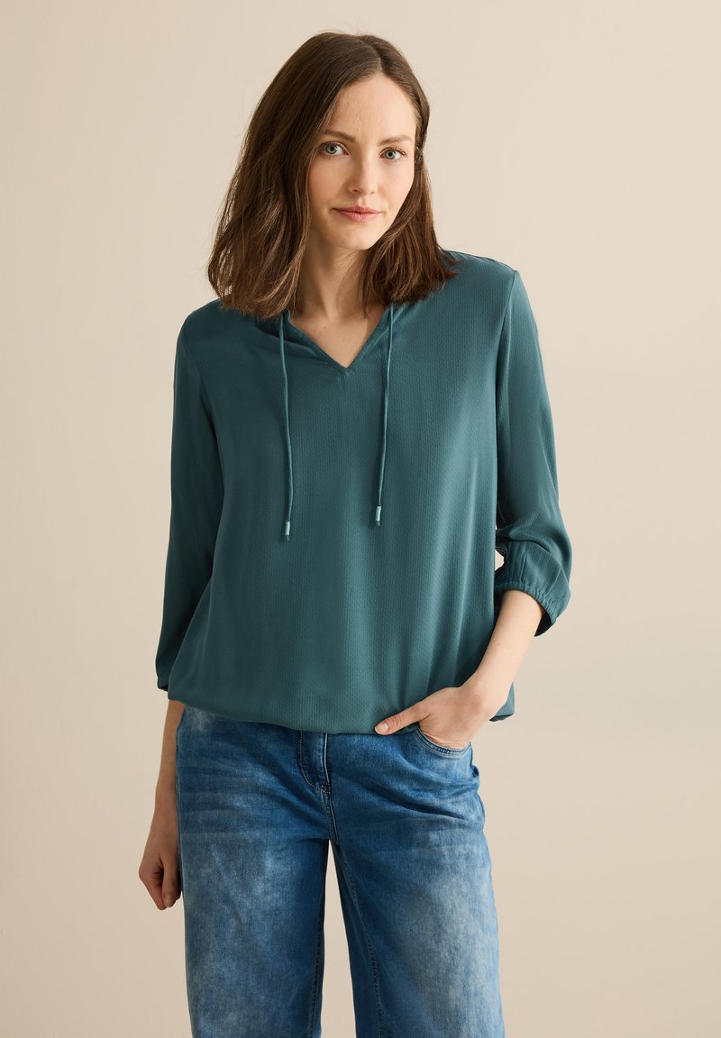 Teal, let bluse med lange ærmer, V-udskæring og snore-detalje. Kombineret med falmede blå jeans. Glat tekstur.
