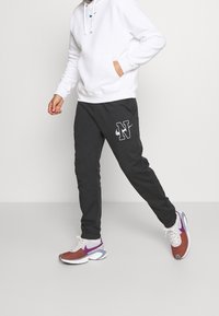 Svarta sweatpants med en vit logotyp på vänster lår, i kombination med en vit hoodie och flerfärgade sportskor.