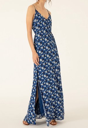 Femme portant une robe maxi bleue sans manches avec imprimé floral blanc et fente latérale, associée à des sandales marron.