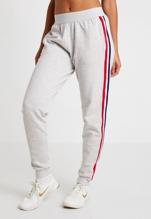 Frau trägt hellgraue Jogginghose mit roten und marineblauen Seitenstreifen und weiße Sportschuhe mit goldenem Swoosh, steht auf weißem Boden.