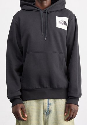Ciemnoszara bluza z kapturem z białym kwadratowym logo The North Face na klatce piersiowej, noszona przez osobę ze swobodnie opuszczonymi rękami i jasnoniebieskimi dżinsami.