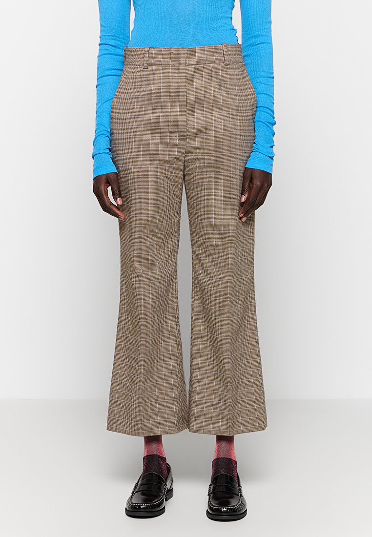 Brązowe spodnie w kratę o rozszerzającym się kroju, z teksturowanej tkaniny, z plisowanym przodem i bocznymi kieszeniami. W połączeniu z czarnymi mokasynami i różowymi skarpetkami.