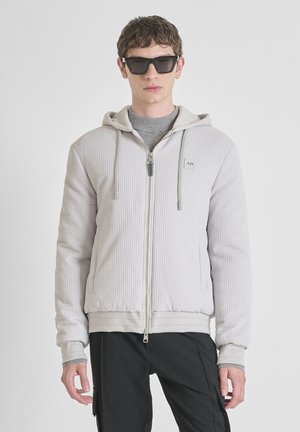 Joven con chaqueta acolchada con capucha de color gris claro sobre una camiseta gris y pantalones cargo negros, con gafas de sol oscuras, de pie contra un fondo blanco.