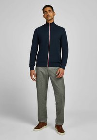 Marine blauwe ritstrui met een witte en rode middenstreep, geribbelde boorden en zoom. Gecombineerd met grijs geruite broek en bruine leren sneakers.